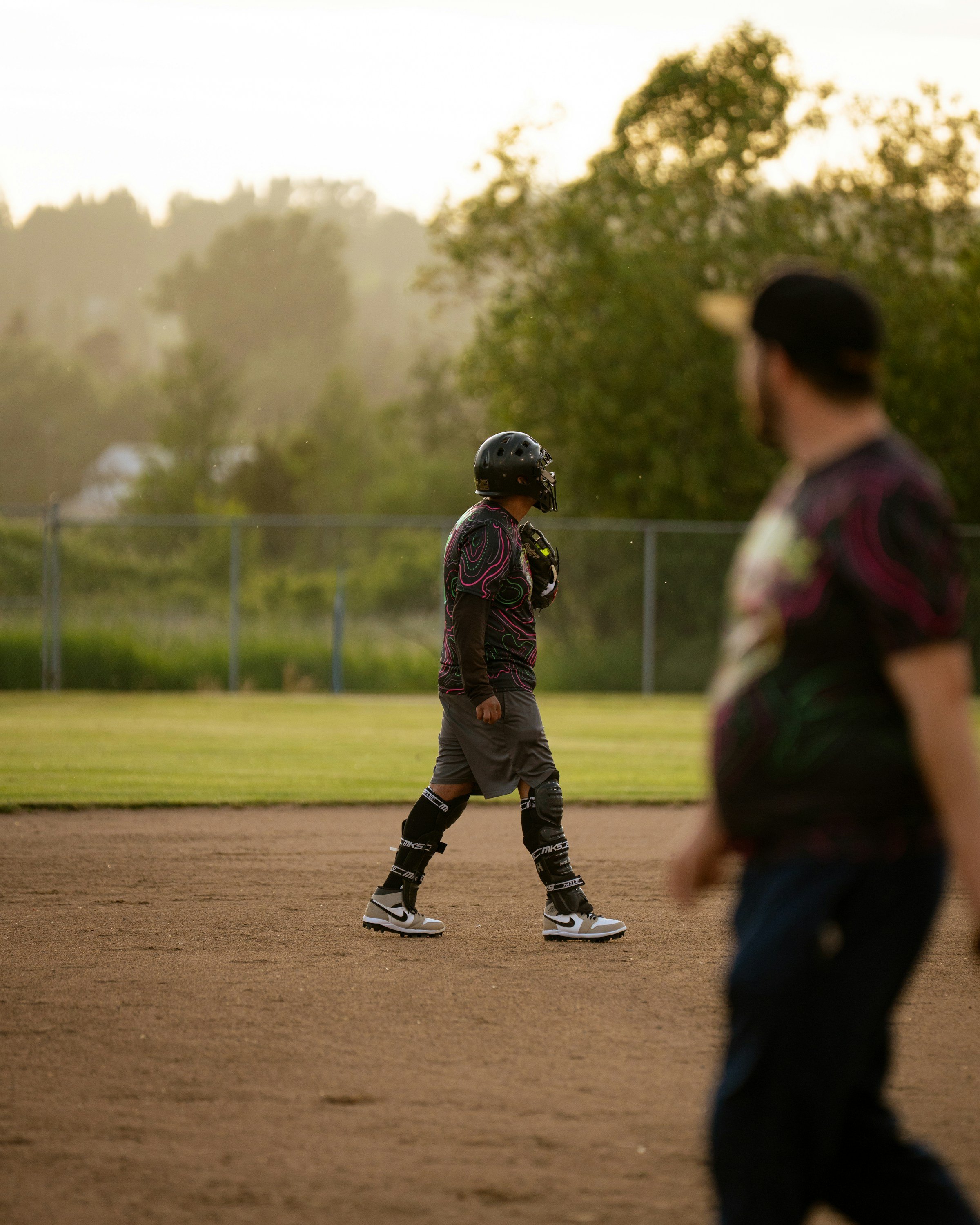 A coach watching a young catcher walk across a baseball field at golden hour